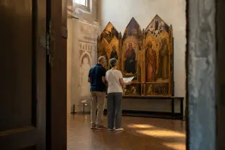 Elderly couple quietly studies a map beside a medieval artwork in a softly lit museum gallery, sunlight streaming through high windows and casting warm reflections on the floor.