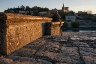 Worn stone pathway beside the Saint-Bénézet Bridge with an elderly man resting his hand on the parapet, bathed in warm golden light.