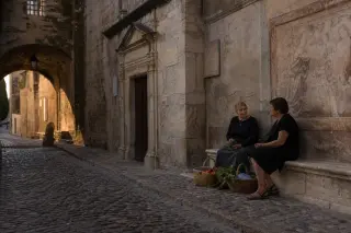 Two people in a quiet cobbled alley of Villeneuve-lès-Avignon, chatting near a stone wall with soft golden sunlight and historic details in the background.