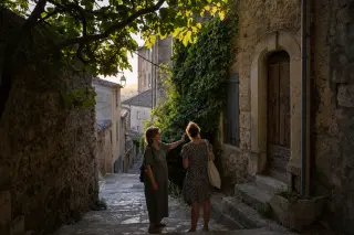 A quiet stone alleyway in Villeneuve-lès-Avignon at golden hour, with a local resident showing a curious visitor an ivy-covered old doorway, both engaged and absorbed, sunlight filtering through leaves.
