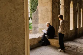 Elderly man sketching ancient inscription in Chartreuse cloister at sunset, teenager observing, natural golden light, intimate documentary angle