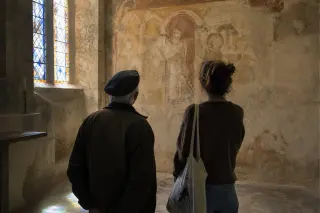 Two people quietly observing a medieval fresco inside a softly lit historic chapel, sunlight casting gentle hues from a stained-glass window onto stone walls and floor.
