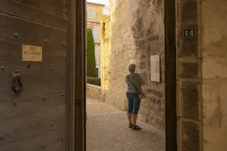 Older woman reading an information plaque in a narrow, sunlit medieval stone passageway, relaxed and immersed in the quiet historical atmosphere.