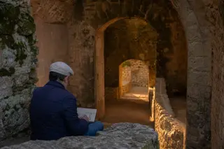 Warm, natural light filters through arched stone inside an old fortress, with a man quietly sketching on a parapet, evoking historical discovery and calm.