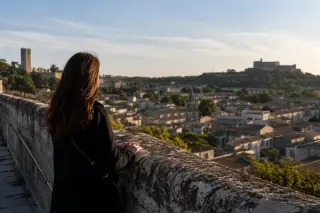Person absorbed in the view from a stone rampart at golden hour, overlooking rooftops with both Tour Philippe-le-Bel and Fort Saint-André faintly visible in the background.