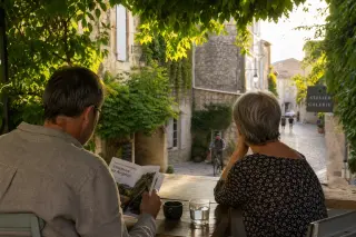 Two visitors quietly enjoying a shaded café terrace along a peaceful stone street in Villeneuve-lès-Avignon at sunset, with a cyclist passing in the background and eco-guide in hand.