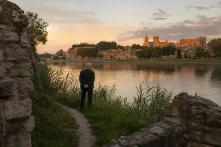 Elderly person quietly observing Avignon from a grassy Rhône riverbank at sunset, with reeds and worn stonework framing the contemplative scene.